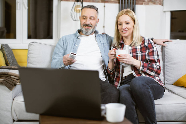 Relaxed married couple sitting on sofa and using laptop - 写真・画像