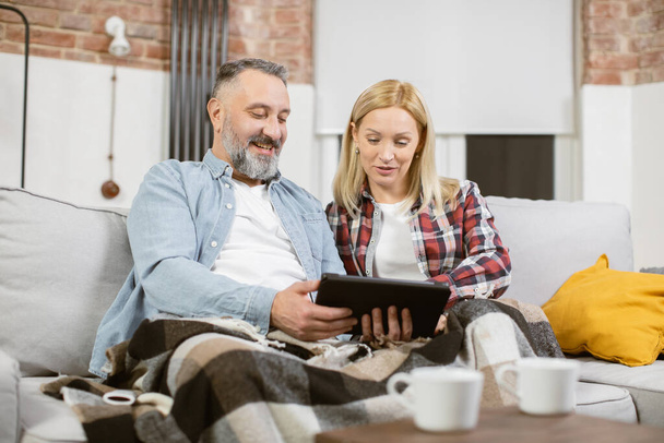 Mature couple sitting on couch with digital tablet in hands - Valokuva, kuva