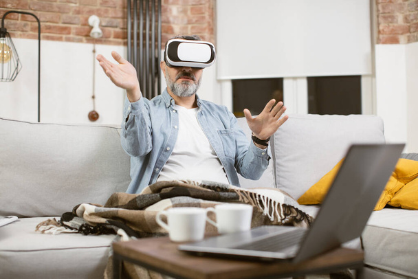Middle aged man sitting on couch in VR headset and gesturing - Photo, Image