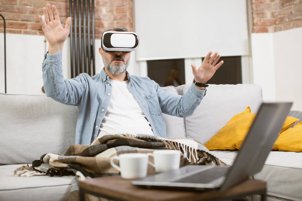 Middle aged man sitting on couch in VR headset and gesturing - Foto, Imagen