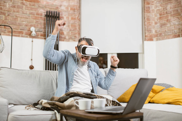 Excited mature man gesturing on couch in VR headset - Photo, Image