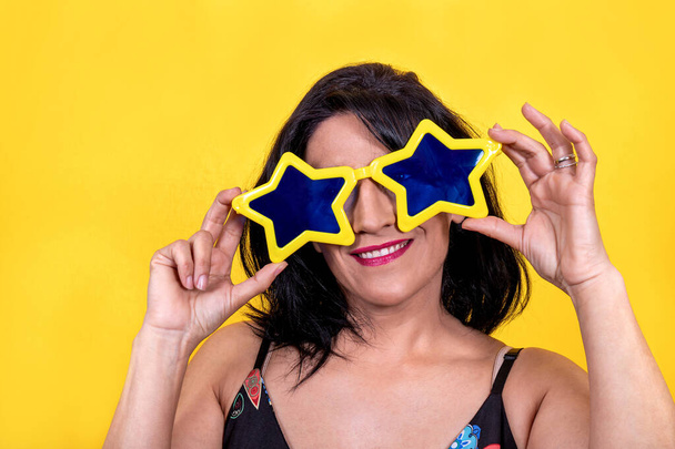 Beautiful woman is wearing yellow star-shaped glasses. Young brunette woman is happy and having fun at the party. - 写真・画像