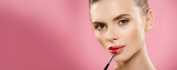 Beauty Concept - Mujer que aplica lápiz labial rojo con fondo de estudio rosa. Hermosa chica hace maquillaje
. - Foto, Imagen