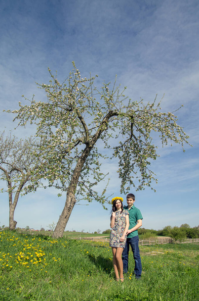 Loving couple near a blossoming tree on a background of spring blue sky. Girl with a wreath of dandelions on her head. - 写真・画像