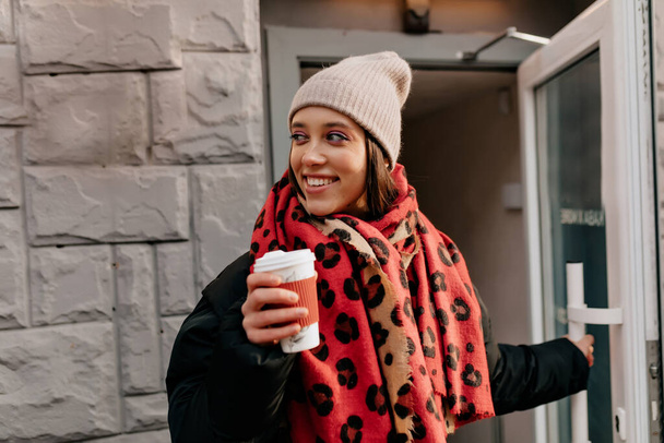 půvabná okouzlující dáma s nádherným úsměvem v pletené čepici a barevnou šálu vycházející z kavárny s kávou jít. Šťastná žena tráví čas ve městě za teplého slunečného dne. Kvalitní fotografie - Fotografie, Obrázek