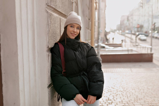 Стильная красотка в бежевой кепке и тёмной куртке позирует на улице на фоне старой европейской улицы. Высокое качество фото - Фото, изображение