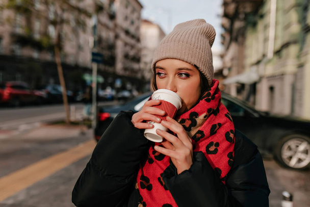 Menina despreocupada com grande maquiagem beber chá na rua ensolarada no centro da cidade. Foto de mulher morena agradável em casaco escuro desfrutando de café. - Foto, Imagem