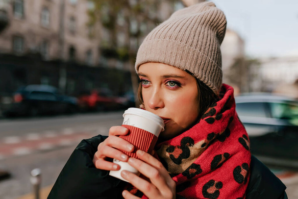 Close up outside photo of beautiful girl with green eyes wearing knitted cap and red scarf enjoying walking in sunny city and drinking coffee. High quality photo - 写真・画像