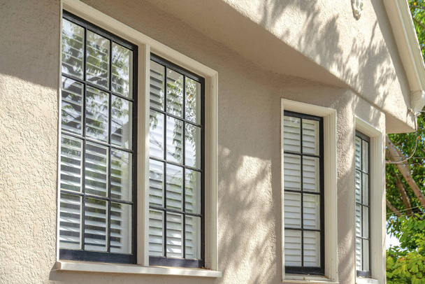 Ventanas de una casa con paredes de hormigón rugoso beige en San Francisco, California. Hay ventanas con rejillas negras y marcos blancos, y un cristal transparente con una vista de las persianas de la ventana en el interior. - Foto, imagen