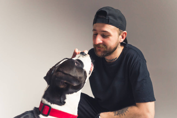 young good-looking bearded and a tattooed man with a cap cuddling his cute dog medium shot gradient background dog concept studio shot  - Fotografie, Obrázek