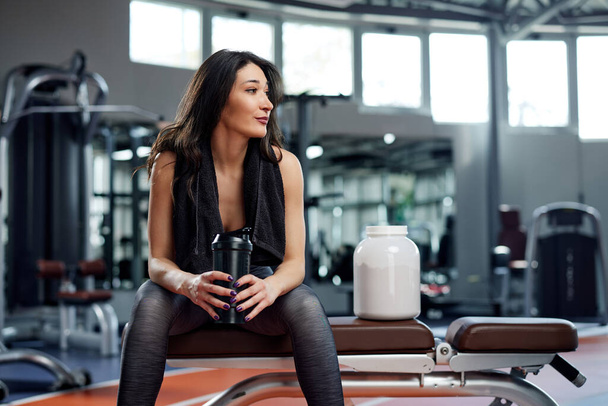 Młoda, aktywna kobieta robi przerwę na siłowni. Siedzi, trzymając proteinowego shake 'a z ręcznikiem na ramionach. - Zdjęcie, obraz