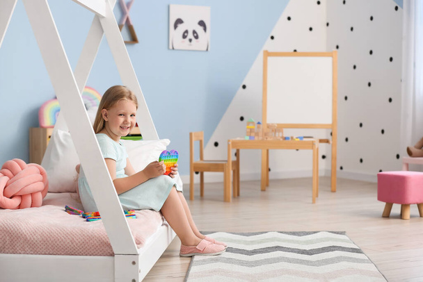 Happy little girl sitting on bed and playing with pop it fidget toy in child room - Photo, image