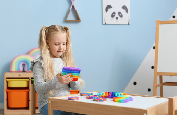 Menina bonita sentada à mesa e brincando com pop multicolorido brinquedo fidget no quarto criança - Foto, Imagem