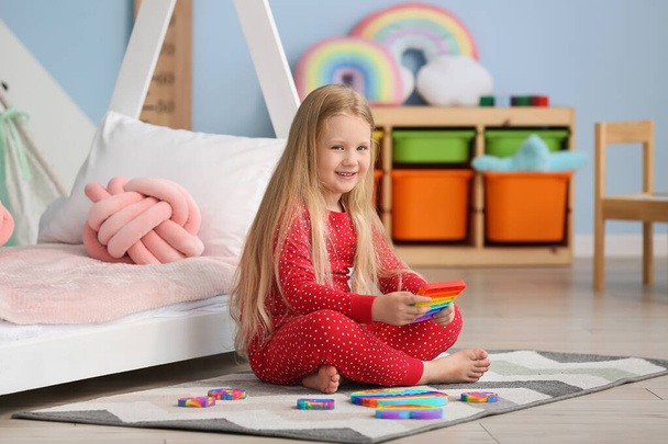 Pretty girl sitting on floor near bed and holding pop it fidget toy in child room - Foto, Bild
