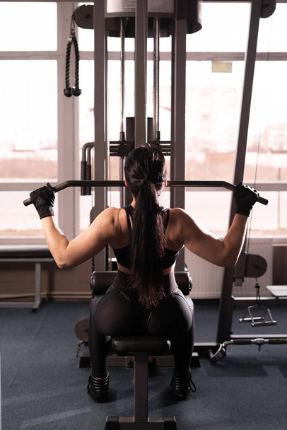 Máquina reductora de hombros. La mujer de fitness está entrenando en el gimnasio. Ejercicio de fuerza superior del cuerpo para la parte superior de la espalda. vista posterior - Foto, Imagen
