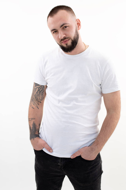Young confident bearded man with short dark hair in white T-shirt, black jeans, standing, thrusting hands into pockets. - Photo, Image
