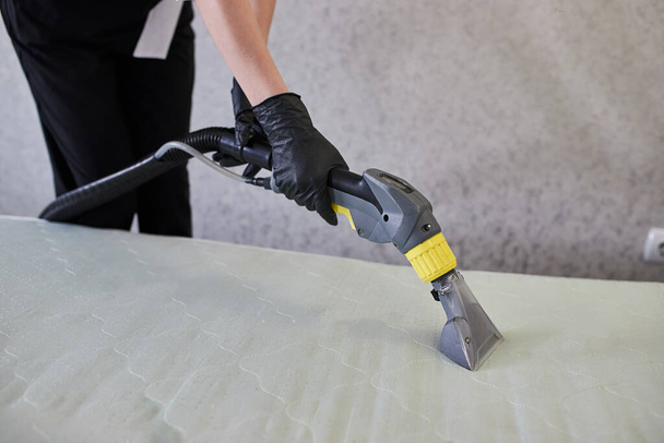 Cleaning service company employee removing dirt from furniture in flat with professional equipment. Female housekeeper arm cleaning the mattress on the bed with washing vacuum cleaner close up - Photo, Image