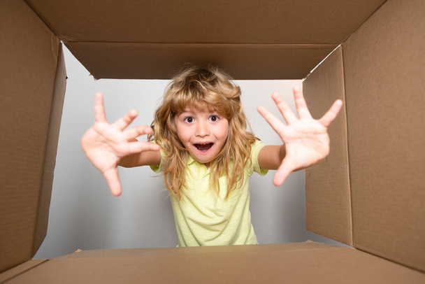 Child opens carton box and pulling out gift from it. Small boy looking in parcel box and happy to receiving a surprise. Young kid showing joy on his face. Unpacking cardboard box for kids. - Zdjęcie, obraz