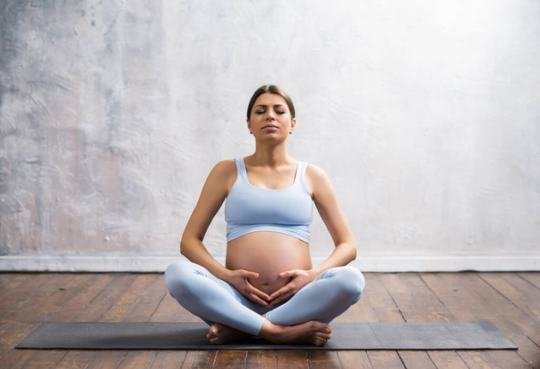 Young pregnant woman doing yoga exercises and meditating at home. Health care, mindfulness, relaxation and wellness concept. - 写真・画像