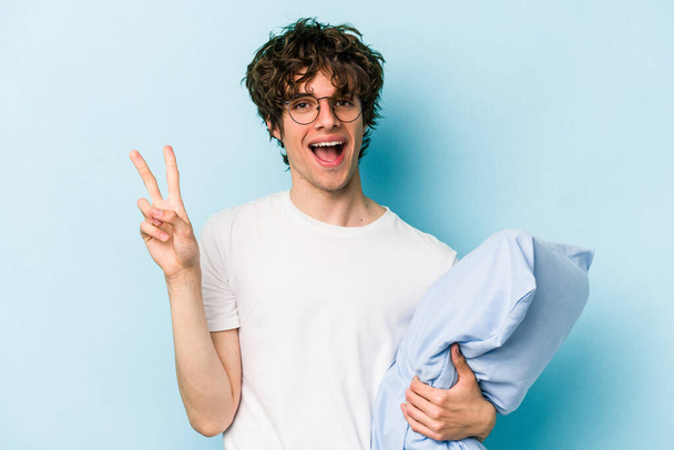 Jonge blanke man draagt een pyjama en houdt kussen geïsoleerd op blauwe achtergrond met nummer twee met vingers. - Foto, afbeelding
