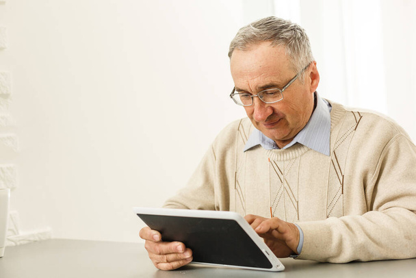 Knappe vrolijke oudere man met behulp van digitale tablet zitten aan de tafel in de woonkamer. Vrijetijdsbesteding, gelukkige pensionering en seniorenlevensstijl - Foto, afbeelding