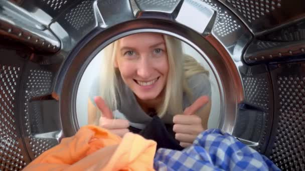 Young beautiful woman doing laundry looks inside washing machine, making normal gesture with smiling hand, shows hand sign to class gesture of approval with happy face. Inside drum. Boondinka. - Felvétel, videó