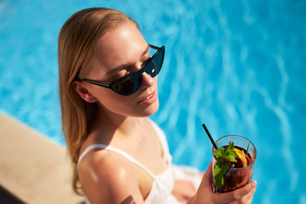 Blonde woman drinks refreshing cocktail sunbathing and sitting near swimming pool at tropical spa. Female in sunglasses enjoys a drink on poolside. Girl chilling in tropical resort on vacation. - Fotó, kép