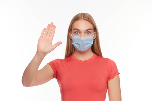 Tıbbi koruyucu maskeli genç bir kadın, stüdyo geçmişinde el hareketleriyle bir dur hareketi gösteriyor. Avuç içini korkuyla dolduran bir kız, karantina önlemleri kavramı. - Fotoğraf, Görsel