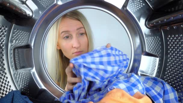 Young Caucasian housewife looks into drum of washing machine, takes out shirt with hole on it. Washing machine ruined clothes, tore them up. Woman is very surprised and upset. Call wizard, warranty - Footage, Video