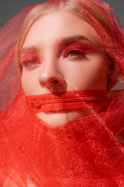 Jovem mulher em pano de tule e zíper vermelho na boca isolado em cinza  - Foto, Imagem