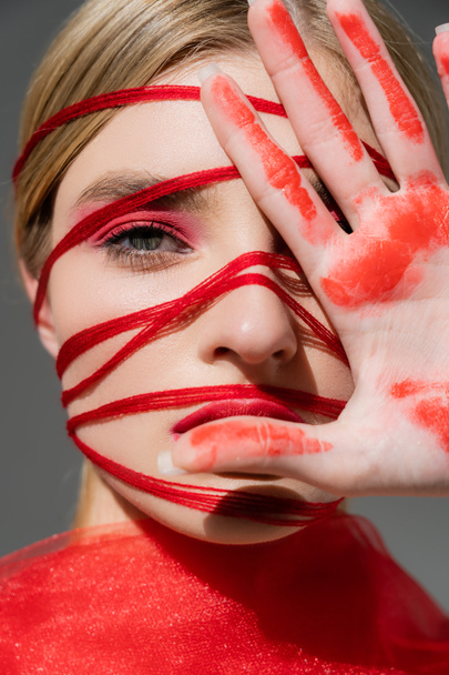 Portrait of woman with red threads on face and paint on hand isolated on grey  - Photo, Image