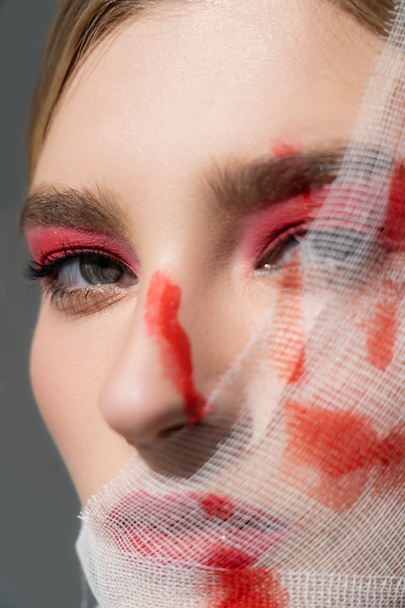 Visão de perto da mulher com tinta vermelha na bandagem médica no rosto olhando para a câmera isolada no cinza  - Foto, Imagem