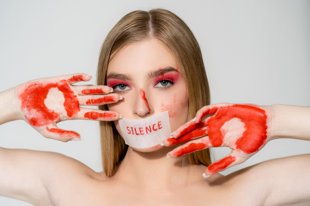 Young woman with paint on hands and lettering silence on paper on mouth isolated on grey  - Photo, Image