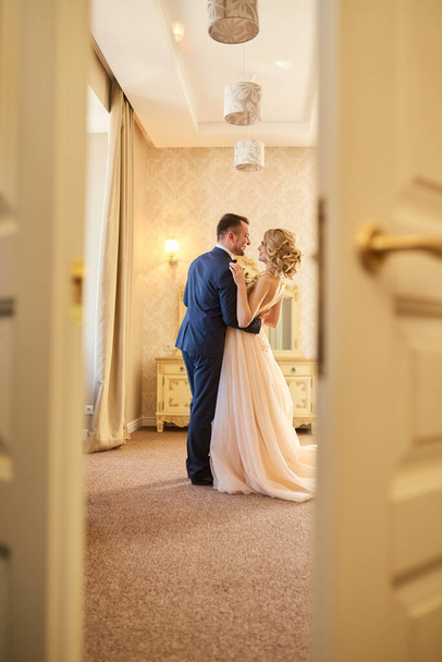 Beautiful couple of young bride and groom in luxurious outfits embracing in elegant interior - Photo, image