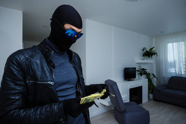 Robber man dressed in black hoodie stands with disguised face and holds a lot of money in his hands, stole a large amount, a thief man stole a TV - 写真・画像