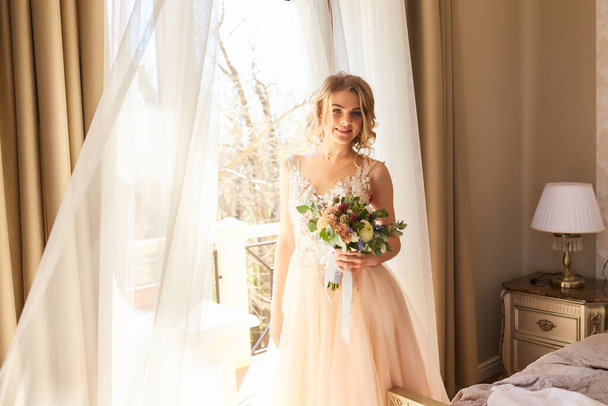 A beautiful bride with a bouquet in hands laughs and poses. Bride smiling near a window - Photo, Image