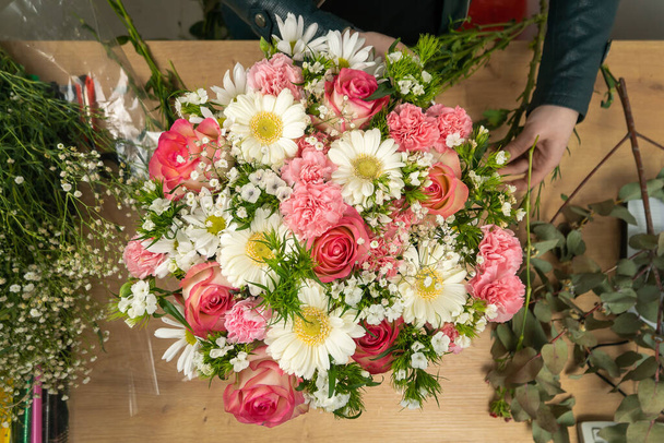 Κάτοψη των χεριών μιας νεαρής ανθοπώλη δημιουργώντας μια όμορφη σύνθεση από ντελικάτα ροζ τριαντάφυλλα, γαρίφαλα και λευκές μαργαρίτες στο τραπέζι - Φωτογραφία, εικόνα