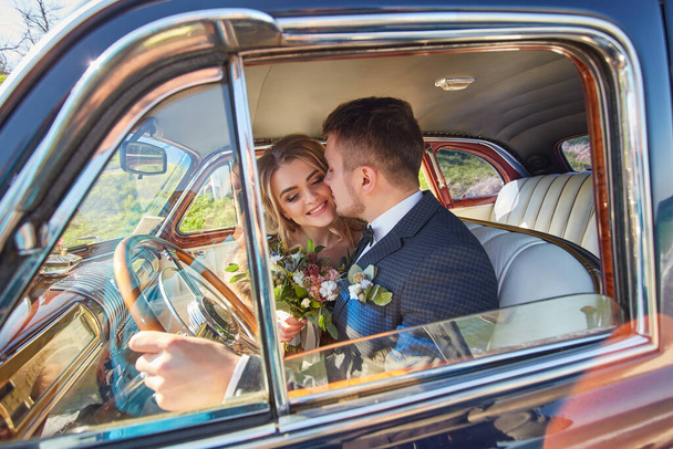 elegante prachtige bruid glimlachend en knappe stijlvolle bruidegom kussen haar in de auto. ongewone luxe bruiloft paar in retro stijl. romantisch moment - Foto, afbeelding