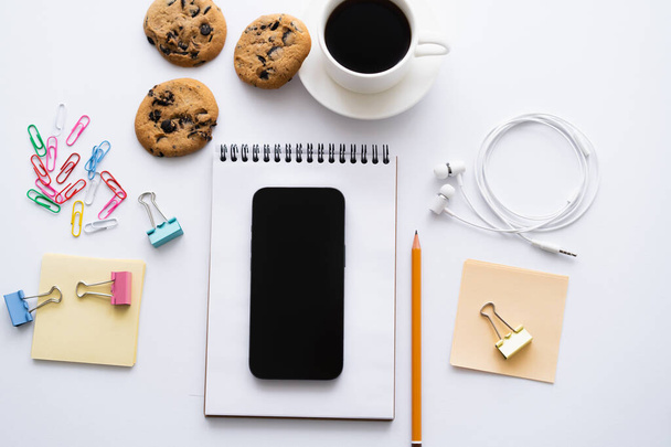 top view of cup of coffee, biscuits and smartphone with blank screen near stationery on white - Фото, изображение