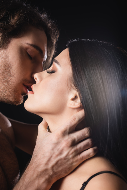 Vue latérale de l'homme passionné touchant le cou de la petite amie brune isolée sur noir  - Photo, image