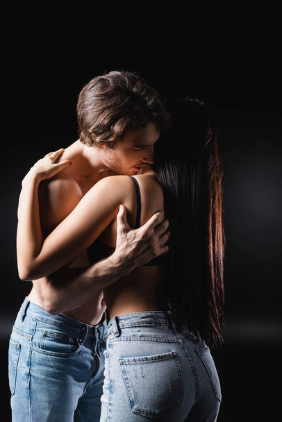 Man in jeans touching brunette girlfriend in bra on black background - Foto, Imagen