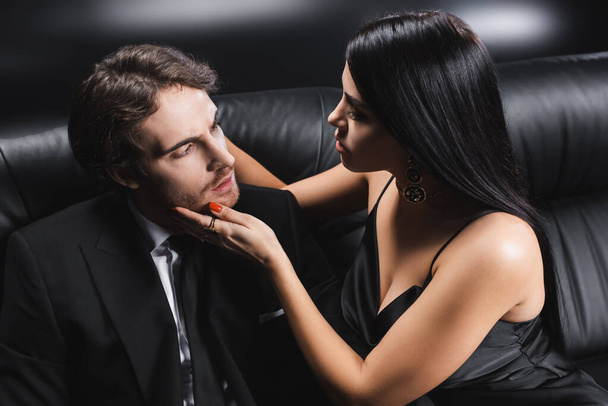 Sexy woman in satin dress touching boyfriend in suit on couch on black background - Fotoğraf, Görsel