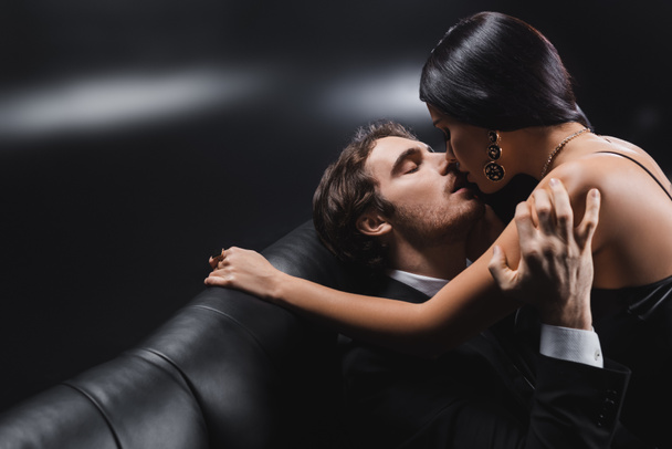 Νεαρός άνδρας με κοστούμι φιλάει μελαχρινή κοπέλα στον καναπέ σε μαύρο φόντο - Φωτογραφία, εικόνα