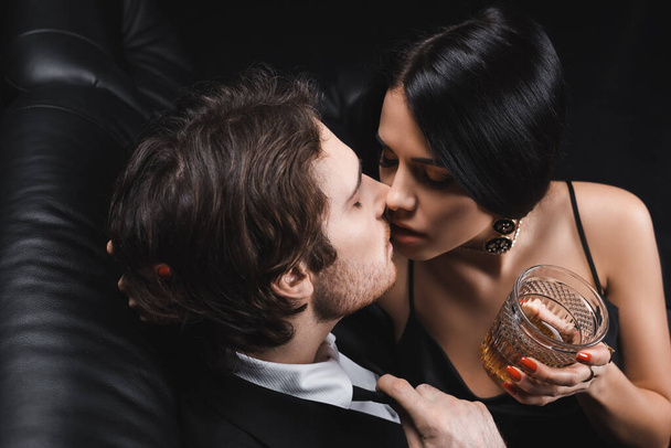 High angle view of brunette woman holding whiskey and kissing boyfriend in suit on couch on black background - Photo, Image