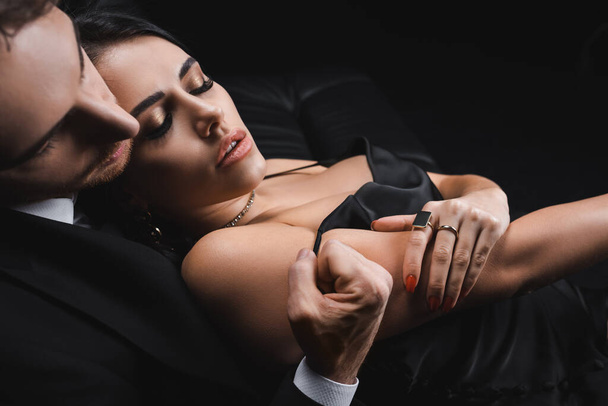 Man in suit undressing sensual woman in silk dress isolated on black  - Фото, изображение