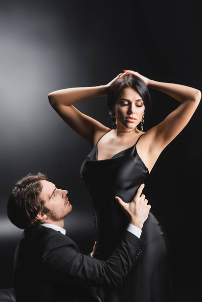 Man in suit touching waist of sexy girlfriend in silk dress on black background - Valokuva, kuva