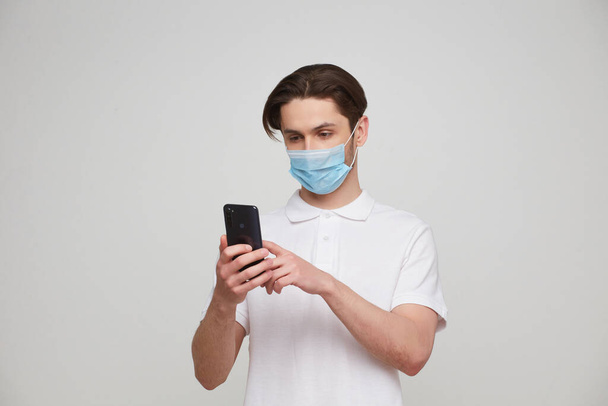 Een Europeaan met een masker gebruikt een telefoon. Conceptuele foto over het thema van de covid-2019 pandemie. Studiofoto - Foto, afbeelding