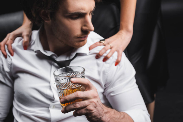 Glass of whiskey in hand of blurred man in shirt sitting near girlfriend on couch isolated on black  - Fotoğraf, Görsel