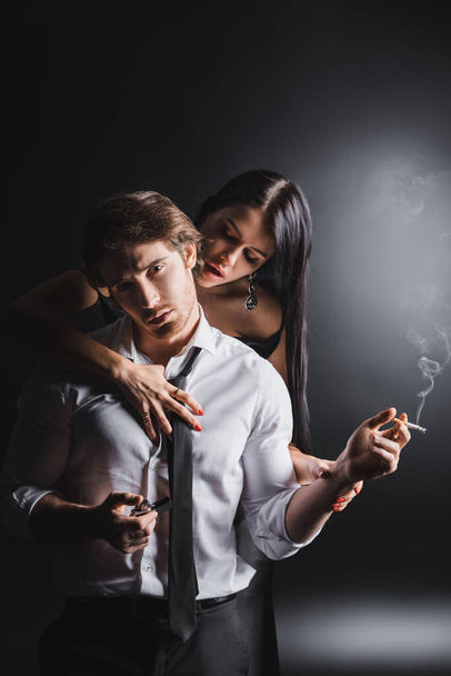 Passionate woman undressing boyfriend in formal wear holding cigarette and lighter on black background - Foto, afbeelding