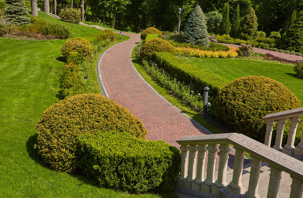 Escaleras con barandillas de piedra balaustres y faroles de hierro en el fondo del parque con un lujoso diseño paisajístico sendero para caminar, césped verde y una variedad de arbustos
. - Foto, imagen